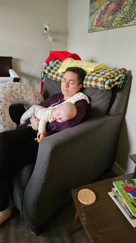 A woman relaxing in a chair, holding a sleeping baby in her arms, with a cozy blanket draped over her shoulder.