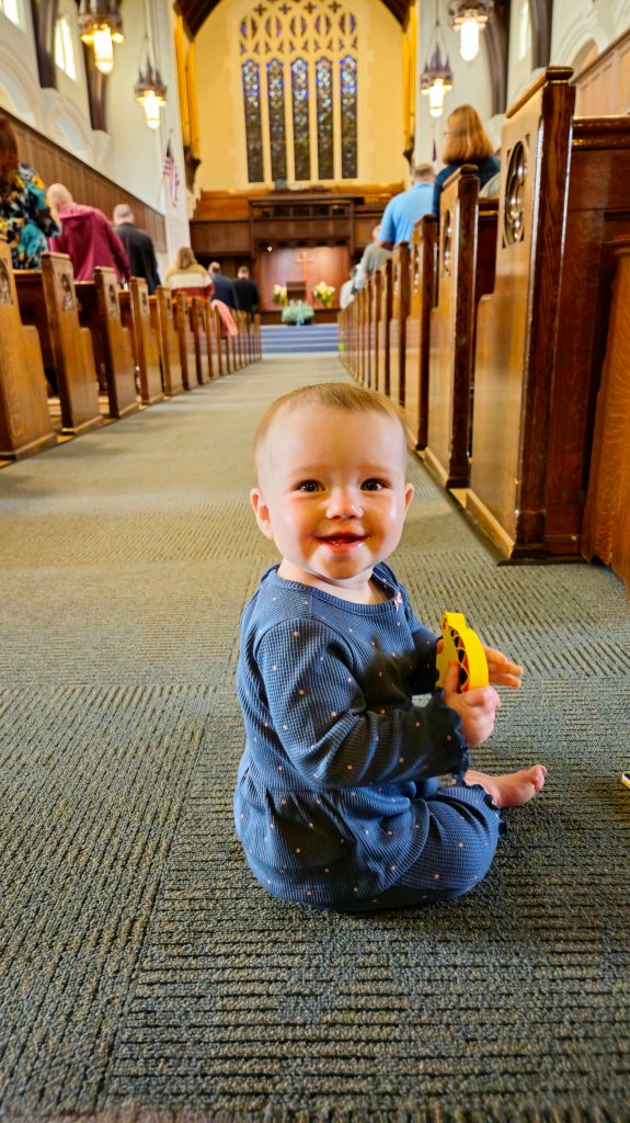 A smiling baby sitting on the carpeted floor of a church, holding a yellow toy, with a view of the church interior in the background.