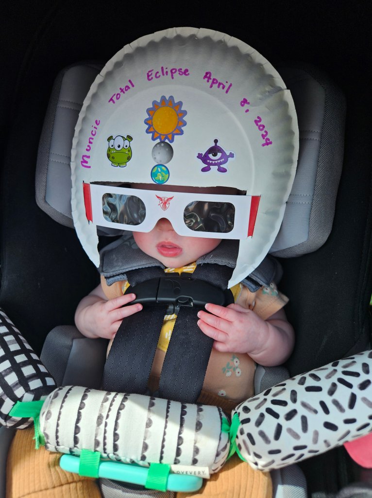 A child wearing eclipse glasses and a decorative paper plate hat inside a car, ready to observe the total solar eclipse.