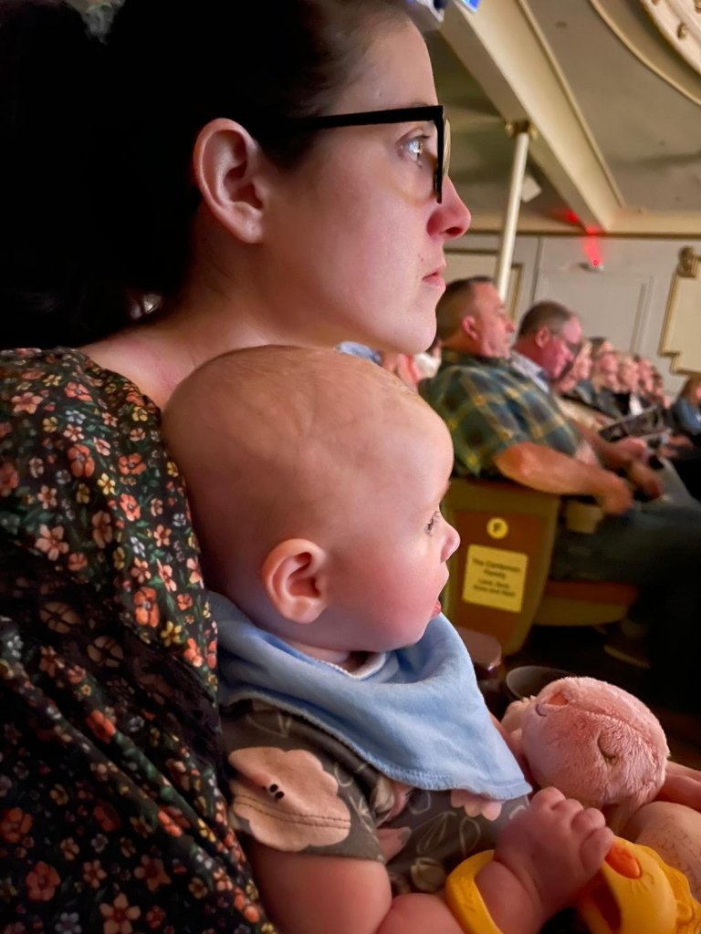 A woman holds a baby close while watching a performance, both appearing engaged in the moment. The audience is visible in the background.